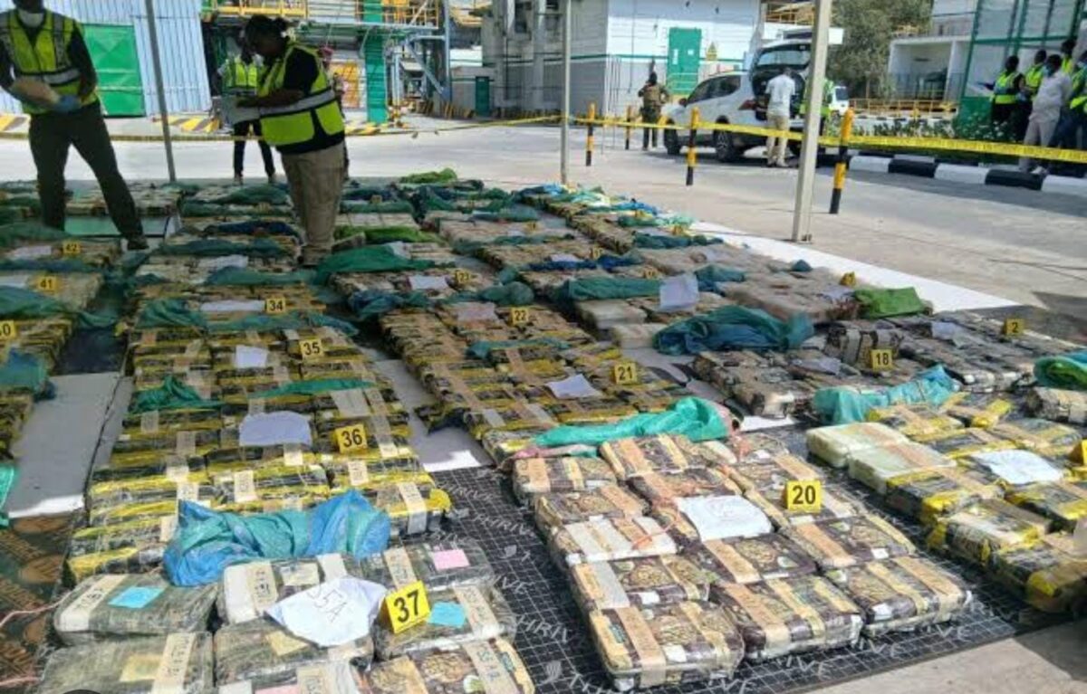 Rows of taped packages and green bags marked with numbered evidence tags lie under a canopy as officials in reflective vests inspect them.