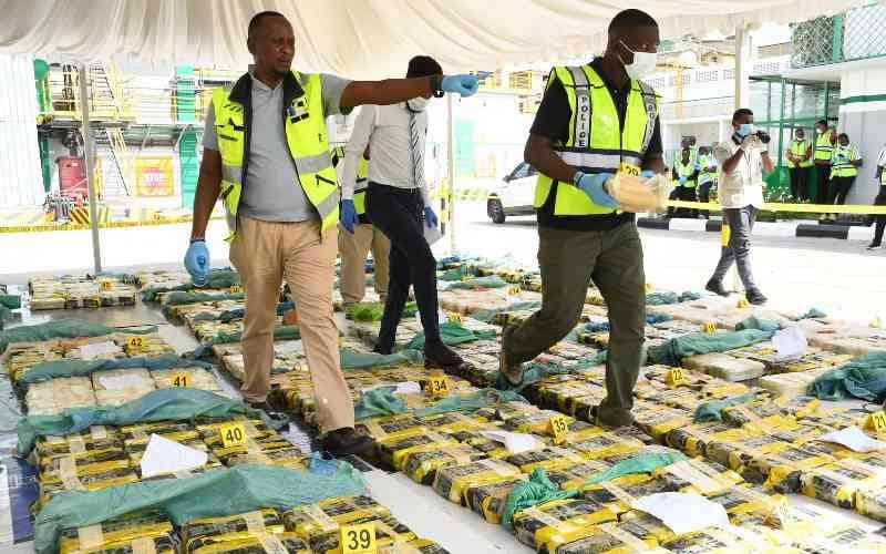 Officials wearing reflective vests, masks and gloves walk among rows of taped packages marked with numbered evidence tags under a canopy.